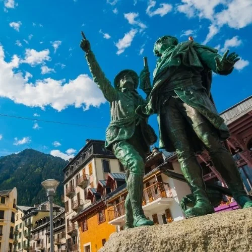 Statue de deux hommes dont l’un pointe vers le ciel, au centre d’un village de montagne.