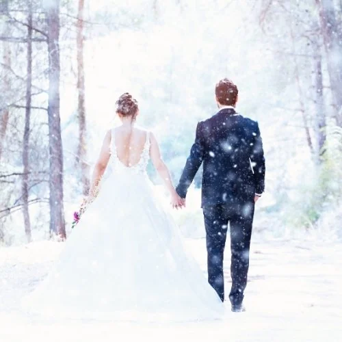 Un couple de mariés marche main dans la main dans une forêt enneigée.