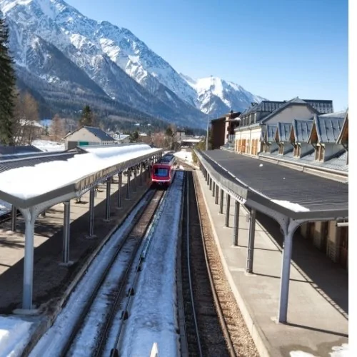 Un train rouge entre les quais d’une gare au pied de montagnes enneigées.
